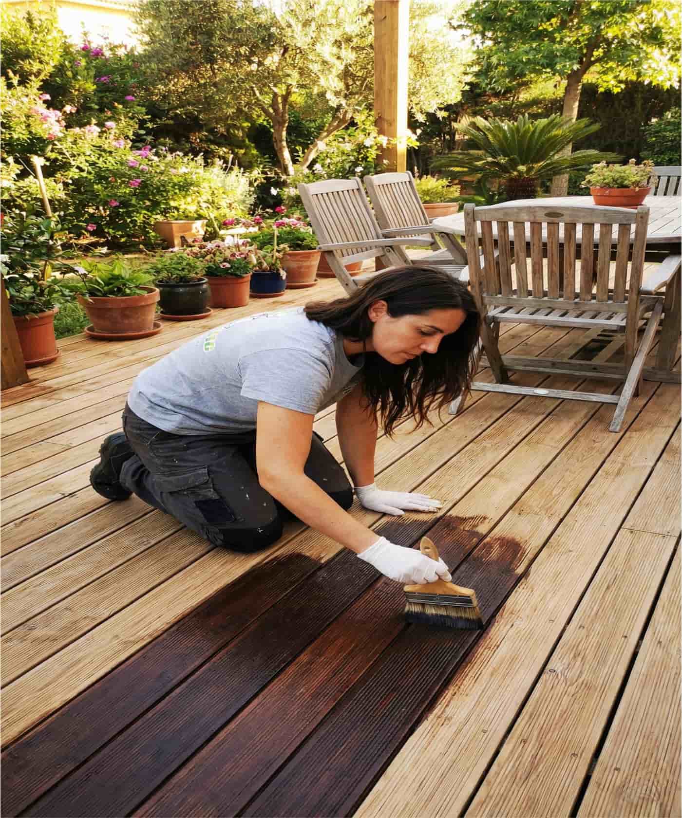 Persona arrodillada aplicando barniz o lasur con brocha sobre una tarima exterior de madera.