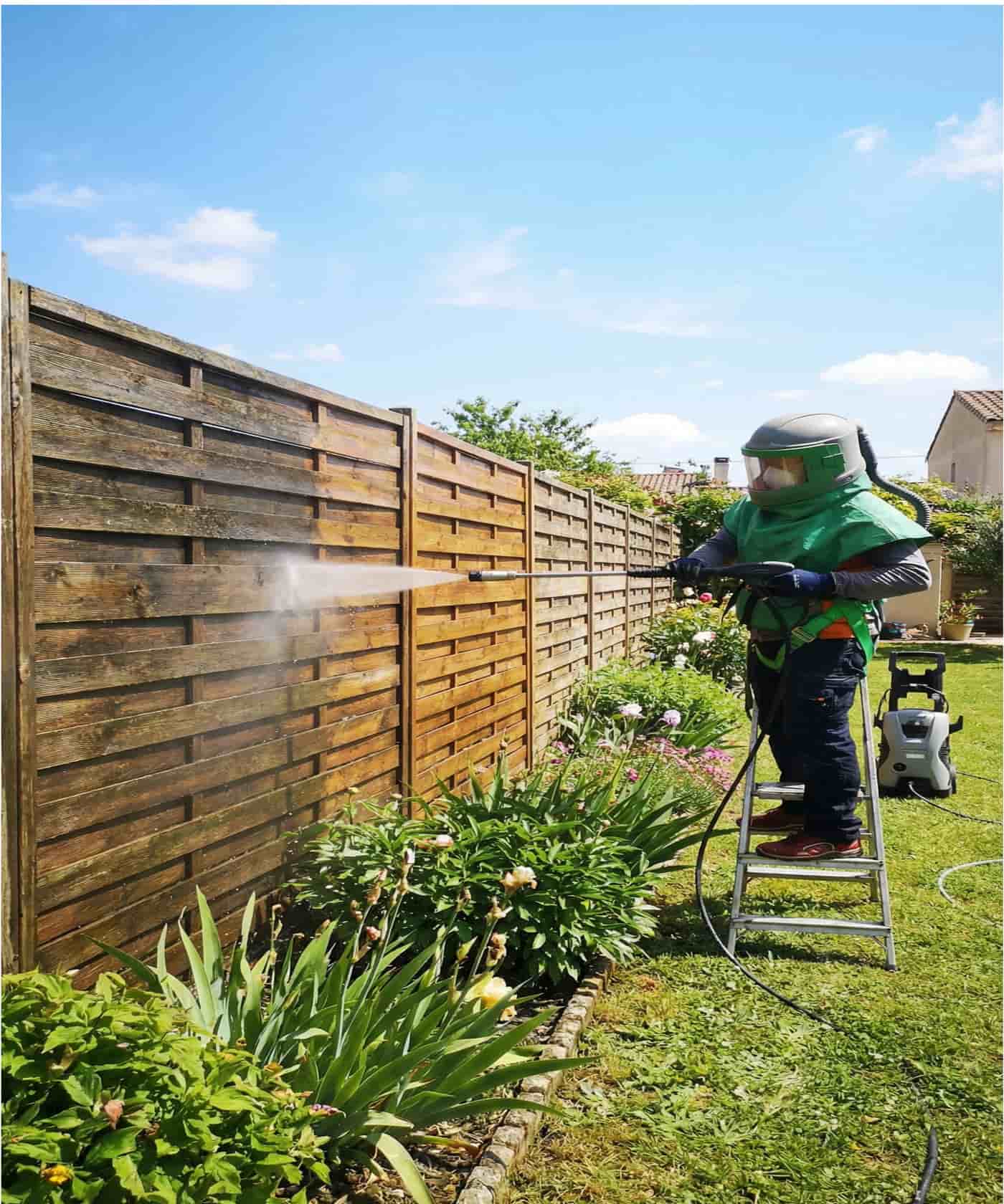 Operario limpiando una valla de madera con hidrolimpiadora desde una escalera baja en el jardín.